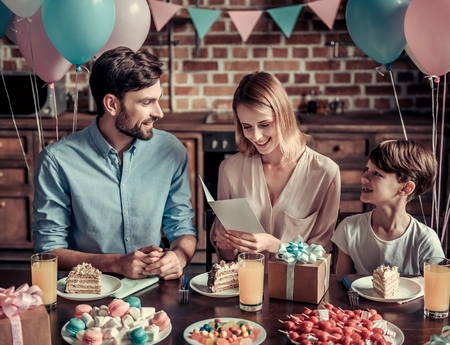 Familie feiert Geburtstag am Tisch, Mutter hält Geburtstagskarte, rosa und pastellfarbene Ballons im Hintergrund