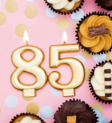 Goldene brennende 85 als Kerze auf Rosa Hintergrund, mit Konfetti und Cupcakes als Design für Einladungskarte zum runden Geburtstag