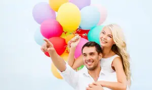 Weiß gekleidetes Paar umarmt sich. Frau hält bunte Helium Luftballons und blickt mit Leichtigkeit in gemeinsame Richtung.