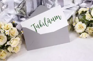 Glückwunschkarte Jubiläum: Eine weiße Jubiläumskarte schaut aus einem grauen Umschlag, auf der Karte stehen Glückwünsche