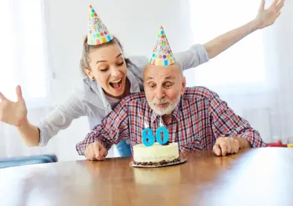Opa bläst Kerze aus, lustige junge Frau dabei, Tragen bunte Partyhüte, als Inspiration für Einladungskarten zum runden Geburtstag