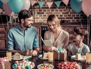 Familie feiert Geburtstag am Tisch, Mutter hält Geburtstagskarte, rosa und pastellfarbene Ballons im Hintergrund