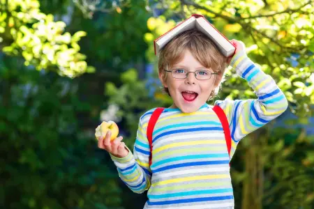 Schulkind Junge zur Einschulung oder Schulstart mit Buch auf dem Kopf und Apfel in der Hand, als Motiv für Einschulungskarten zum Schulbeginn