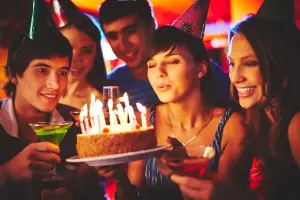 Teenager feiern Geburtstag, Mädchen bläst Kerzen auf Kuchen aus, bunte Lichter im Hintergrund, Motiv für Einladungskarte zum Geburtstag
