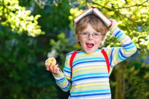 Schulkind Junge zur Einschulung oder Schulstart mit Buch auf dem Kopf und Apfel in der Hand, als Motiv für Einschulungskarten zum Schulbeginn