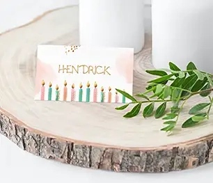 Schöne Platzkarte mit Namen "Hendrick" und bunten Kerzen als Motiv, als Tischdekoration mit weißen Kerzen im Hintergrund