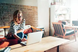 Teenager gestaltet Einladungskarten für ihren Geburtstag am Laptop, sitzt auf cooler Couch