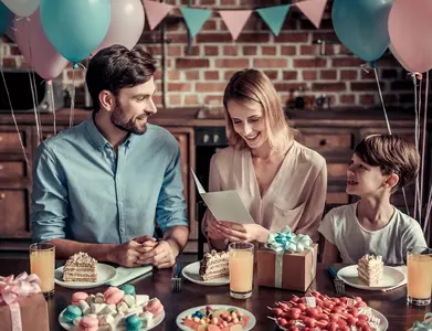 Familie feiert Geburtstag am Tisch, Mutter hält Geburtstagskarte, rosa und pastellfarbene Ballons im Hintergrund