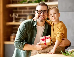 Ein Papa hält ein Vatertagsgeschenk in der Hand und umarmt dabei seinen Sohn. Beide sind sehr lustig und fröhlich